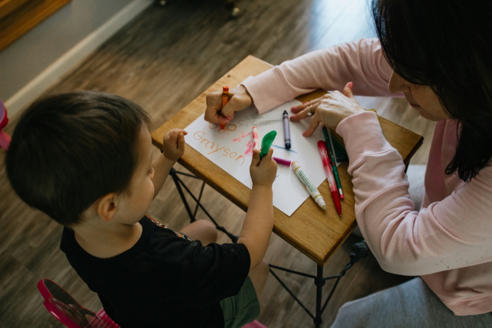 An adult and a child drawing with crayons at a small table, seen from above