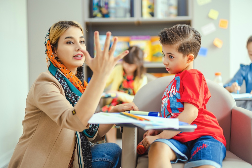 An adult and a child sitting face to face at a table, the adult holding up an open hand as the child watches