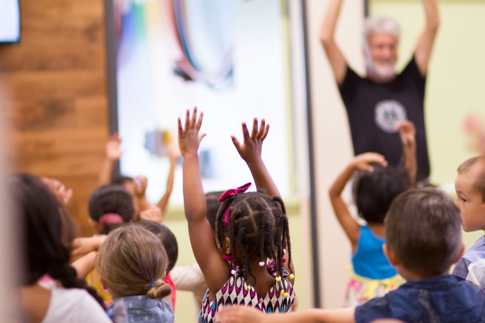Young children with their arms raised, following an adult leading a movement activity