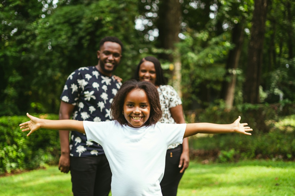 A child standing outdoors with arms outstretched, two adults smiling behind